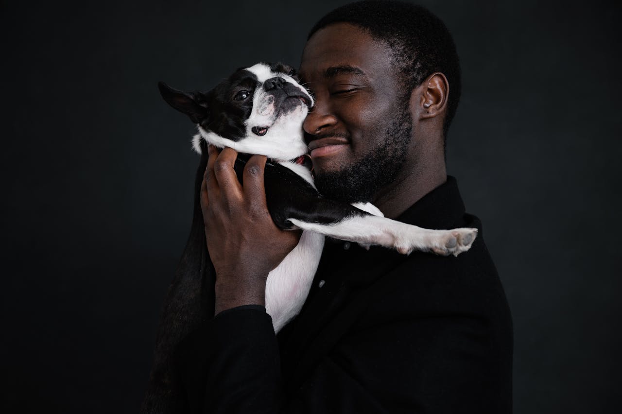 Side view of African American unshaved male in black jacket embracing cute Boston Terrier with closed eyes against black background